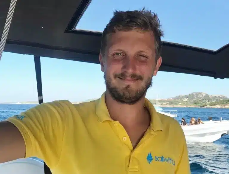 Ein Mann in einem gelben Poloshirt mit Logo lächelt während eines Segeltörns auf einem Boot mit klarem blauen Himmel und Küstenlandschaft im Hintergrund.