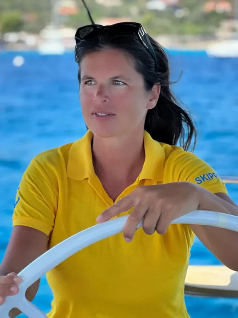 Eine Frau in einem gelben Hemd und mit Sonnenbrille auf dem Kopf steuert ein Boot auf einem Segeltörn, blickt mit konzentriertem Gesichtsausdruck nach vorne, im Hintergrund das blaue Meer.